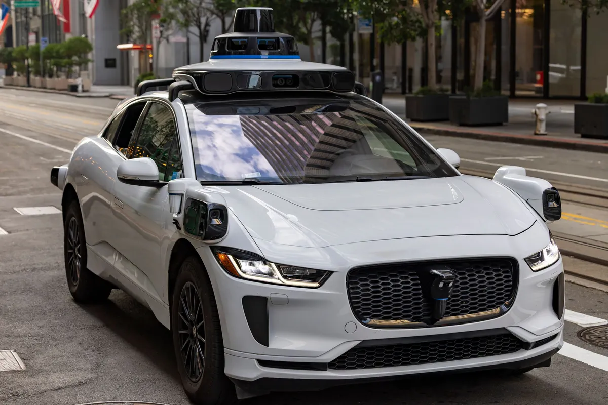A white autonomous vehicle on a city street, buildings reflected in its bodywork.