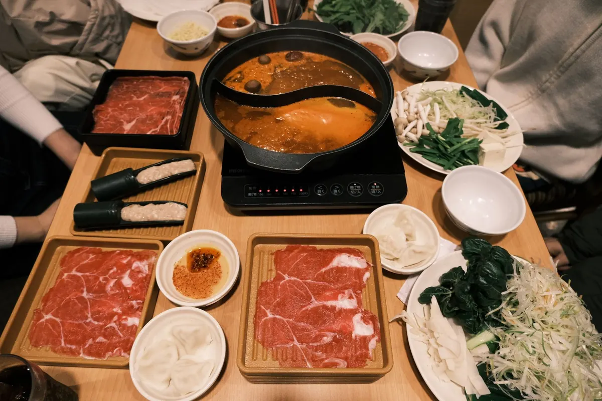 A hotpot table set with a divided broth pot and plates of sliced raw meat and vegetables