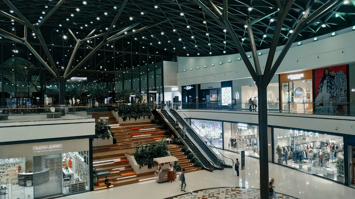The interior of a large modern shopping mall with escalators and planting between the shopfronts